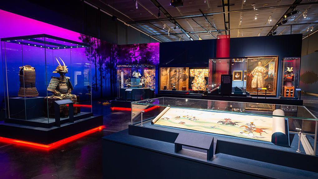 Parte de la muestra de la exposici&oacute;n &ldquo;Samur&aacute;is&rdquo; en el Museo Brit&aacute;nico. En la imagen se aprecia, en primer plano y detr&aacute;s de una vitrina, un documento ilustrado con figuras de personas cabalgando. A la izquierda se observa una armadura, y al fondo se distinguen cuadros y otras im&aacute;genes, aunque no con claridad.