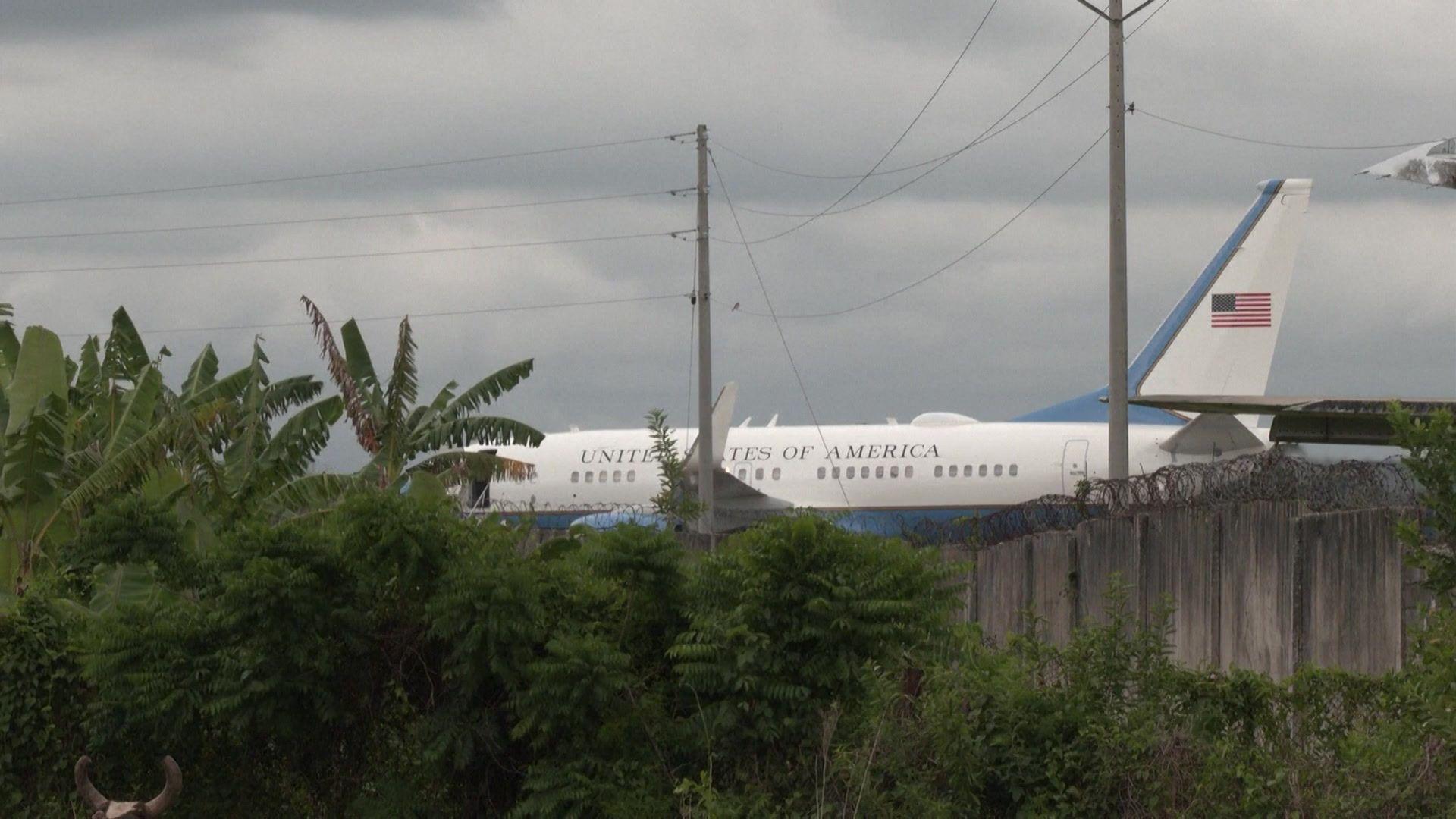 Un avi&oacute;n blanco con letras azules que dicen "Estados Unidos de Am&eacute;rica", rodeado por vegetaci&oacute;n. Tambi&eacute;n se ve un muro con un alambrado de p&uacute;as.