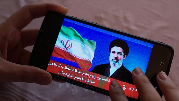 Una mujer sostiene un tel&eacute;fono mientras que en su pantalla se ve una imagen del l&iacute;der supremo de Ir&aacute;n.