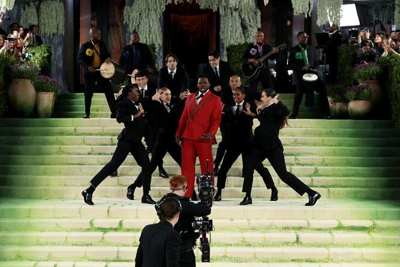 Grupo de personas baila y act&uacute;a en una escalinata verde durante un evento de gala; una persona en el centro viste traje rojo, rodeada de bailarines de negro, con m&uacute;sicos y c&aacute;maras al fondo.