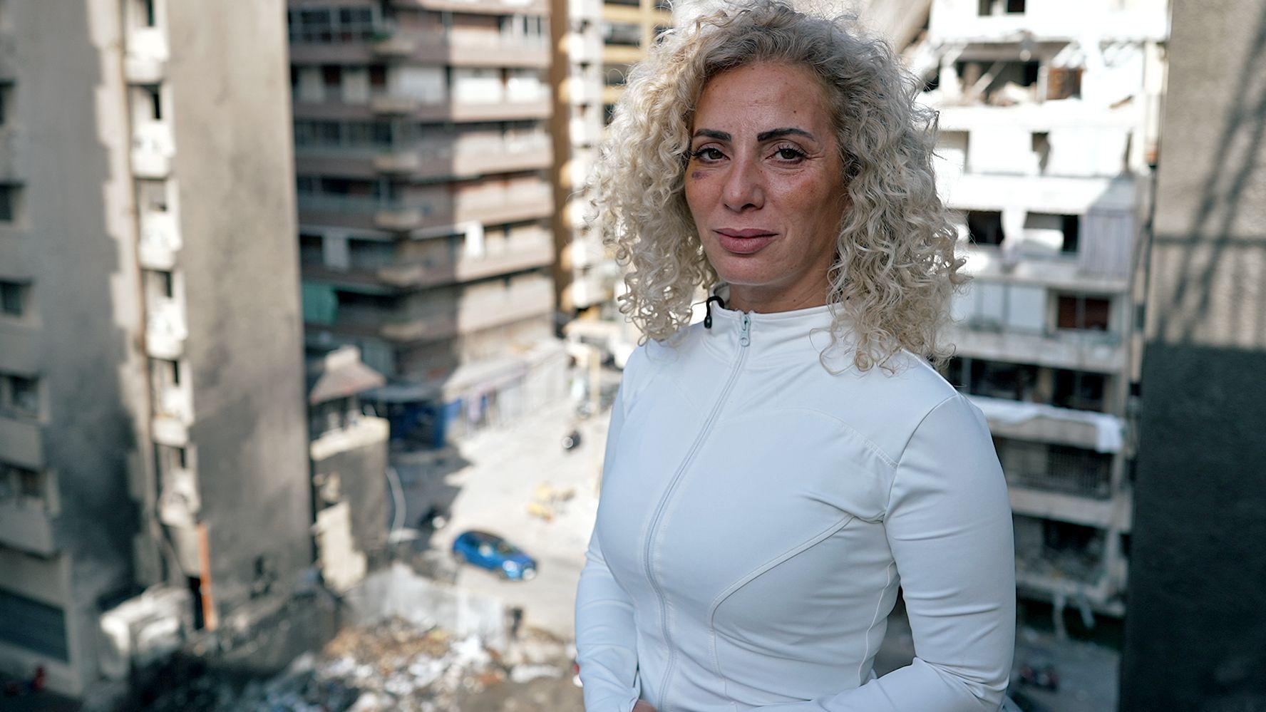 Una mujer parada en un balc&oacute;n, con moretones en la cara, desde donde se mira la destrucci&oacute;n causada por una bomba.