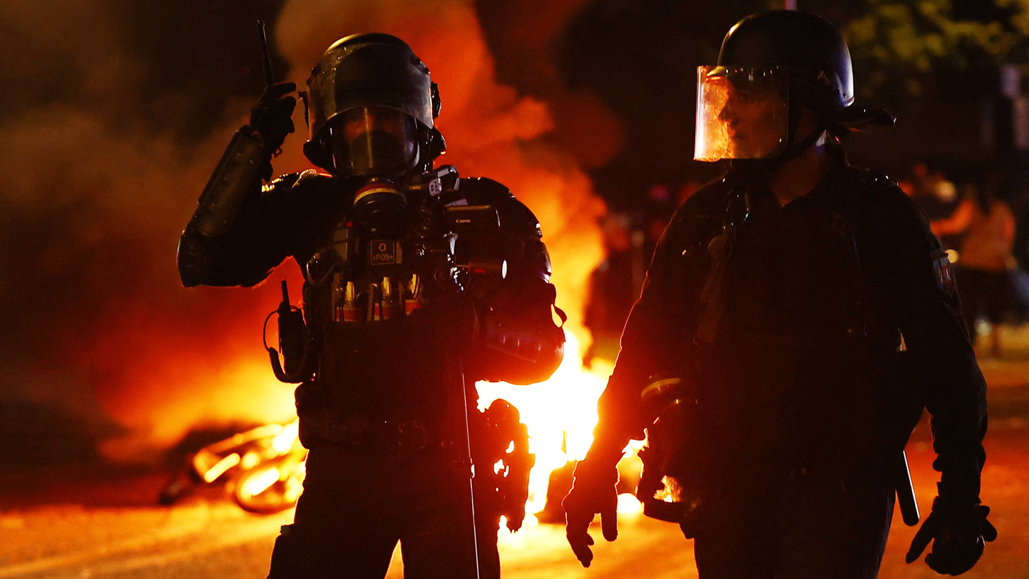 Dos polic&iacute;as, con guantes, cascos y mascarillas de pl&aacute;stico protectoras, caminan por una calle y en el fondo se fuego. 