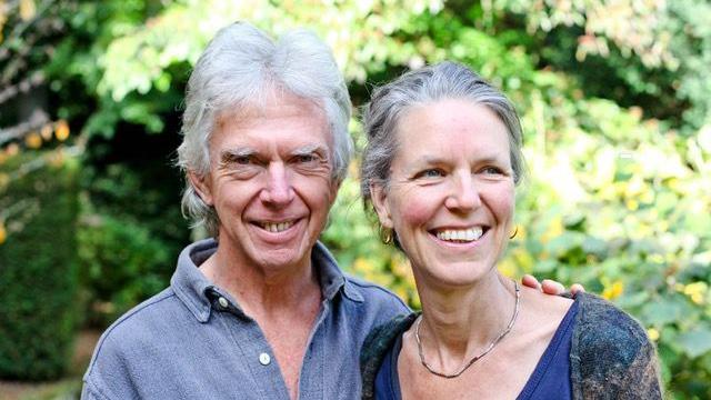 Foto de una mujer y su marido sonriendo. El marido tiene la mano sobre el hombro de su mujer. Los dos van vestidos de azul. Ambos tienen el pelo canoso. Detr&aacute;s de ellos se ven &aacute;rboles y hojas. 