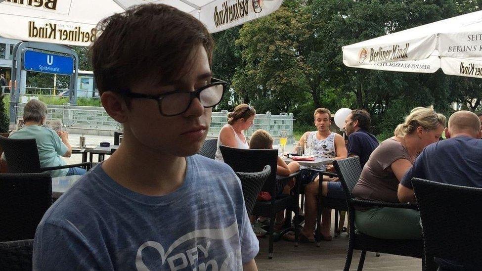 Thomas Parfett, un joven que lleva una camiseta azul descolorida y gafas de montura negra. Est&aacute; sentado en una zona de asientos al aire libre con sombrillas.