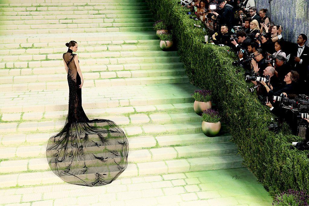 Una persona posa sola en una amplia escalinata verde durante un evento de gala, con un vestido negro con larga cola transparente, mientras fot&oacute;grafos capturan im&aacute;genes desde un lateral.