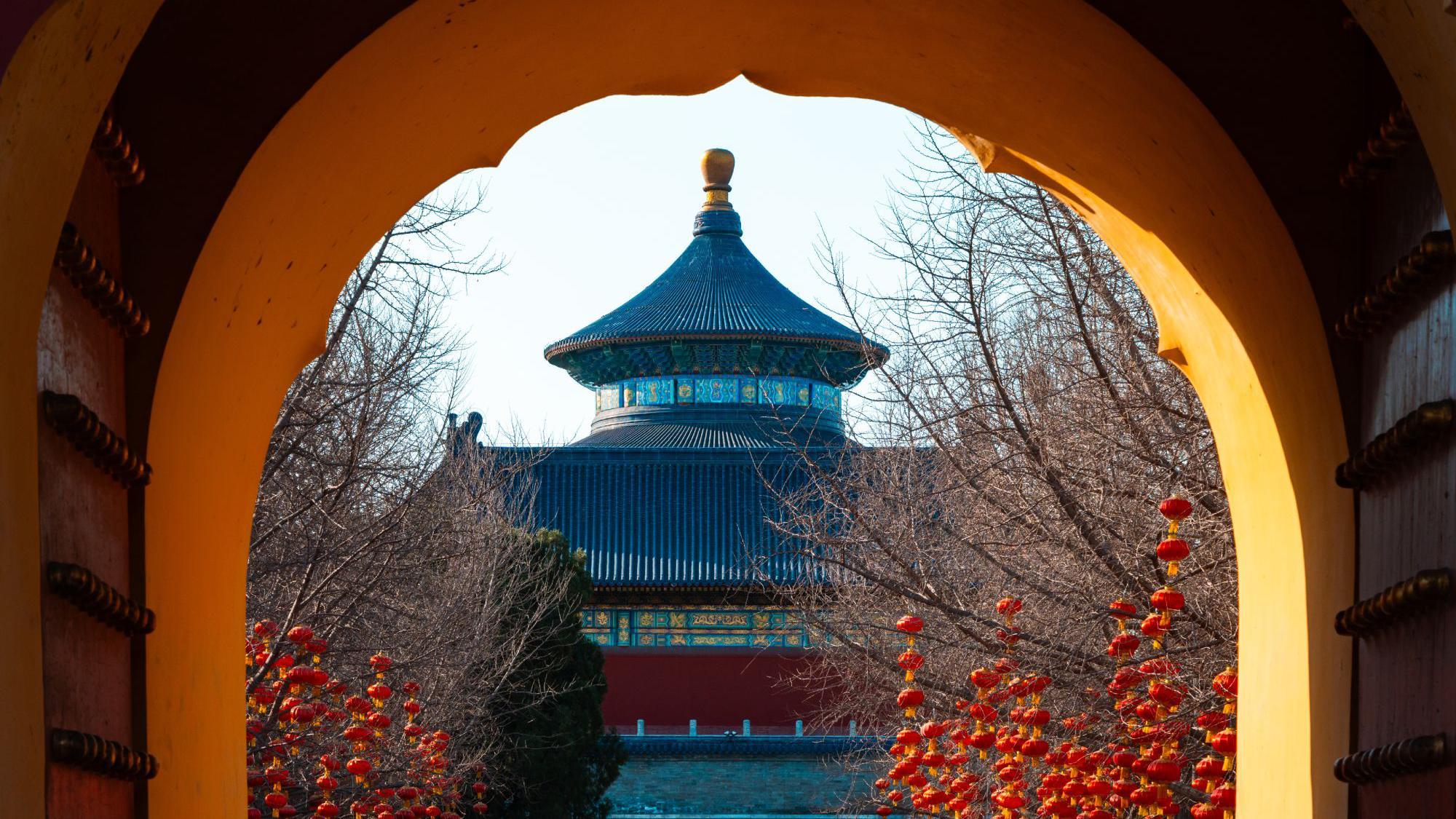 Una vista a trav&eacute;s de un portal hist&oacute;rico, contemplando los ornamentados edificios de un templo en tonos azules, rojos y dorados, con numerosas linternas chinas colgando de los &aacute;rboles desnudos en primer plano.