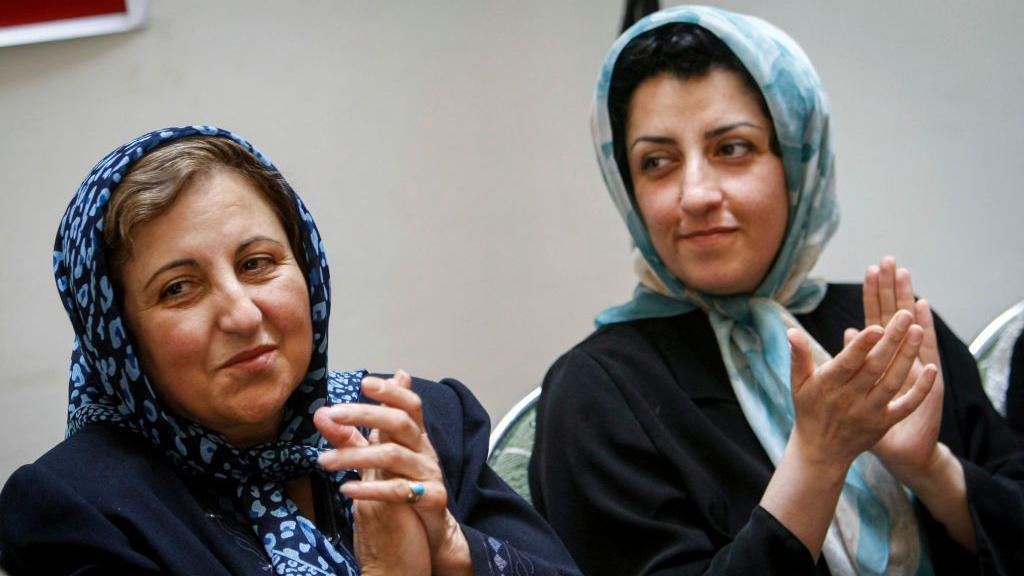 Shirin Ebadi y Narges Mohammadi aplauden durante una conferencia de prensa. Ebadi lleva un pa&ntilde;uelo estampado en azul oscuro, con algo de cabello visible, mientras que Mohammadi lleva un pa&ntilde;uelo estampado en azul claro, con parte de su cabello visible, y una chaqueta negra.