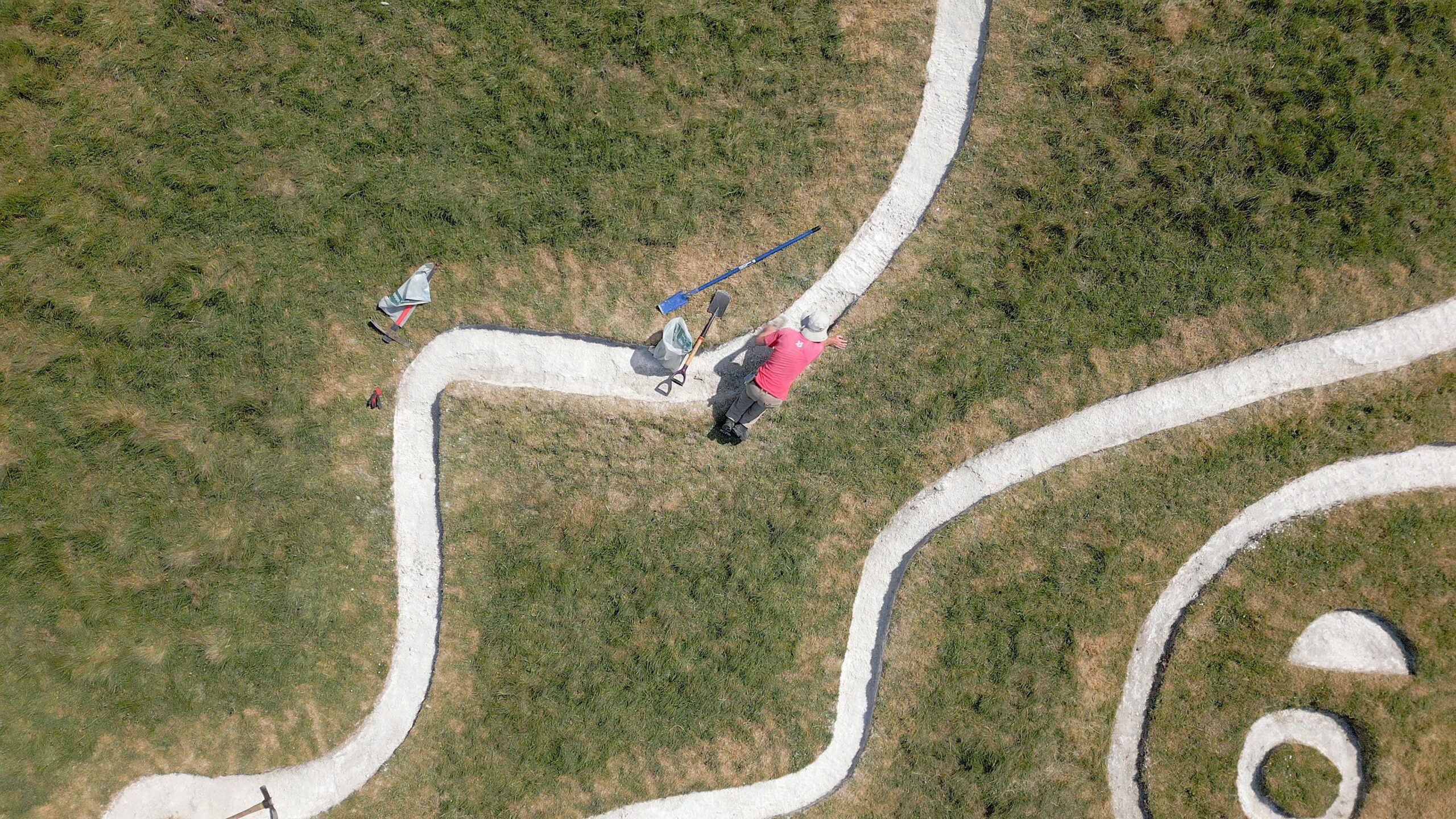 Un hombre con una camiseta roja y un sombrero se ve desde arriba. Est&aacute; a cuatro patas sobre una l&iacute;nea de tiza blanca, con una pala y una bolsa de pl&aacute;stico a su lado. 