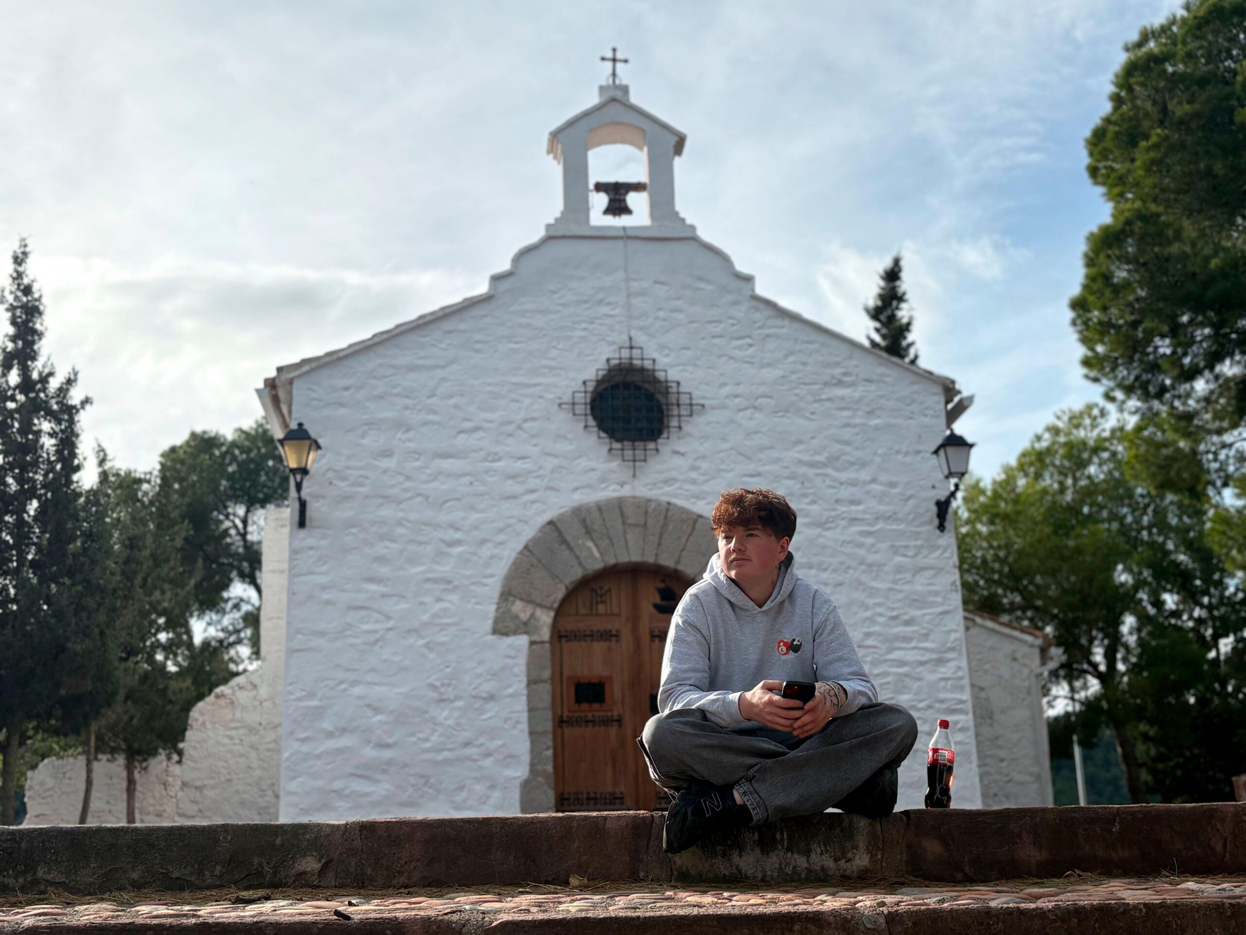 Alex Batty est&aacute; sentado con las piernas cruzadas sobre un muro bajo de piedra, sosteniendo un tel&eacute;fono m&oacute;vil, con una botella de pl&aacute;stico para bebidas colocada a su lado.