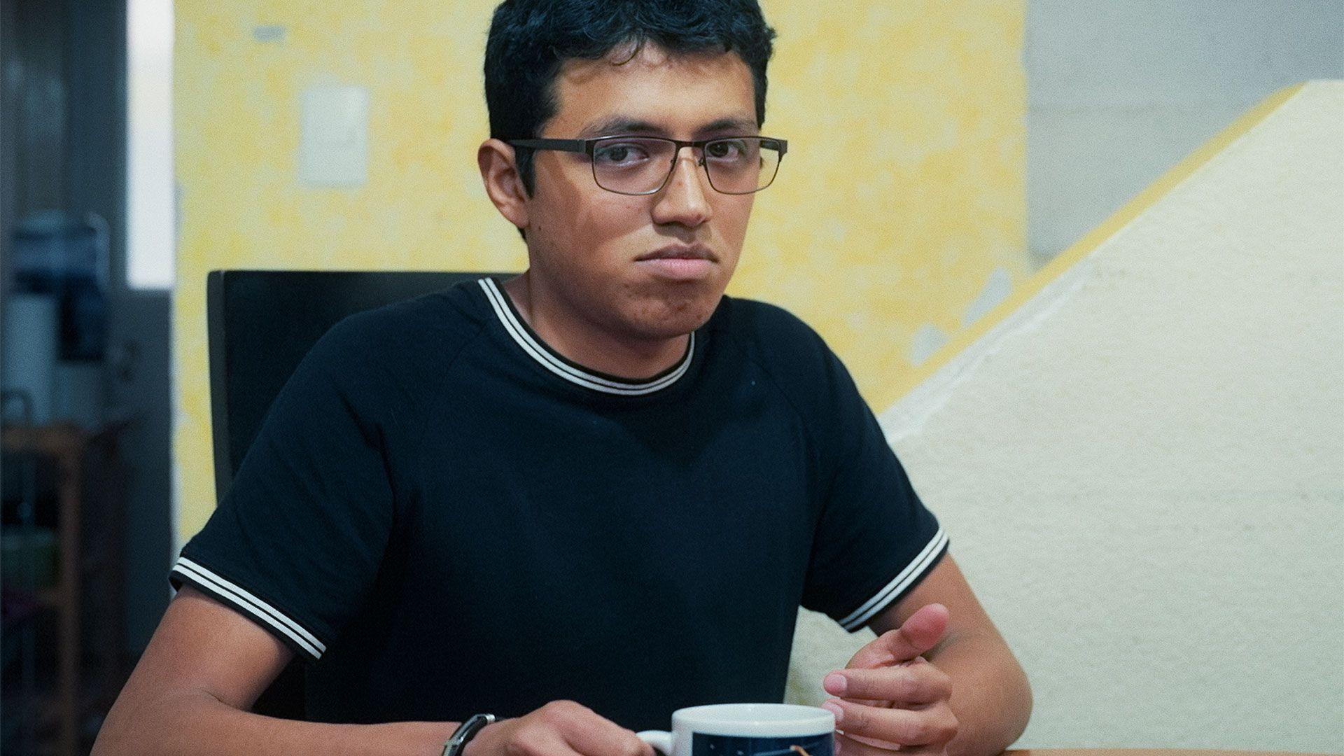 Julian lleva puesta una camiseta azul marino y blanca, y sostiene una taza de t&eacute;; lleva anteojos y tiene el pelo oscuro.