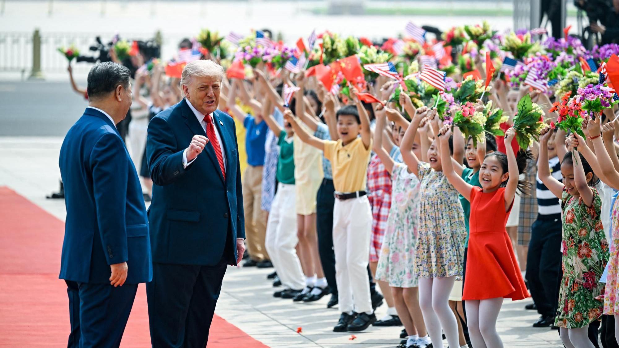 El presidente Trump se sit&uacute;a junto al presidente Xi con el pu&ntilde;o en alto, mientras ambos caminan por una alfombra roja flanqueada por filas de ni&ntilde;os que vitorean y ondean flores, as&iacute; como banderas chinas y estadounidenses.