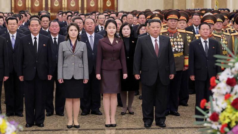 Ri Sol Ju, vestida con un traje de chaqueta gris y  falda, junto a su hija, quien viste un traje similar morado y al lado el l&iacute;der coreano durante un homenaje al fundador del pa&iacute;s.