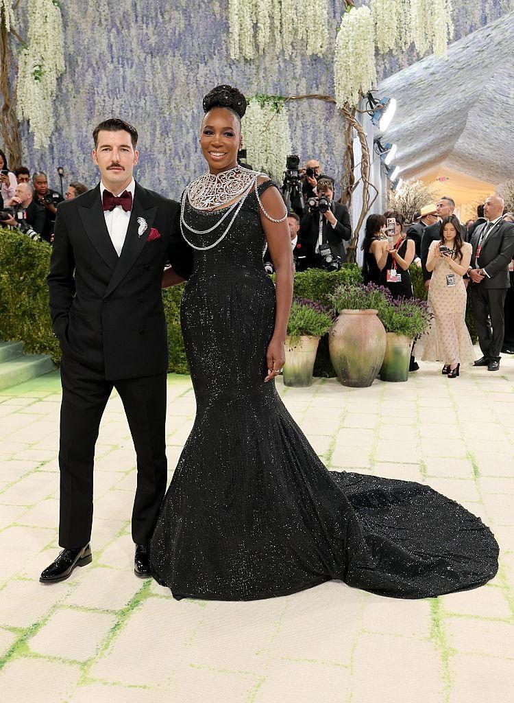 Un hombre y una mujer posan en una alfombra verde durante un evento formal; &eacute;l viste traje negro con pajarita y ella un vestido largo negro con joyer&iacute;a. Hay fot&oacute;grafos y decoraci&oacute;n floral de fondo.