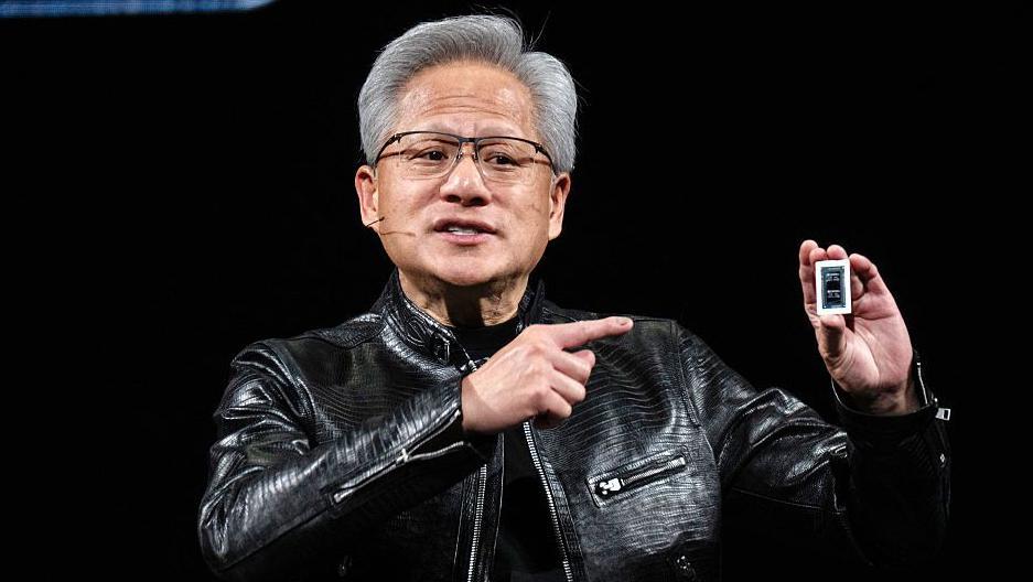 Jensen Huang sostiene un chip de computadora en la mano y lo se&ntilde;ala. Viste de negro y lleva una chaqueta de cuero negra.            