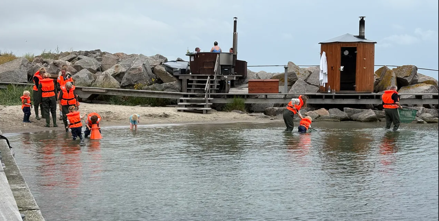 Varias personas, algunas con chalecos salvavidas naranjas, se adentran en aguas poco profundas con redes junto a una orilla rocosa, donde tambi&eacute;n se ven una sauna de madera y una peque&ntilde;a construcci&oacute;n sobre una plataforma.