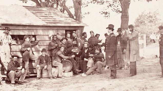 Un grupo de soldados de la guerra de secesi&oacute;n est&aacute; bebiendo frente una casa de madera. 