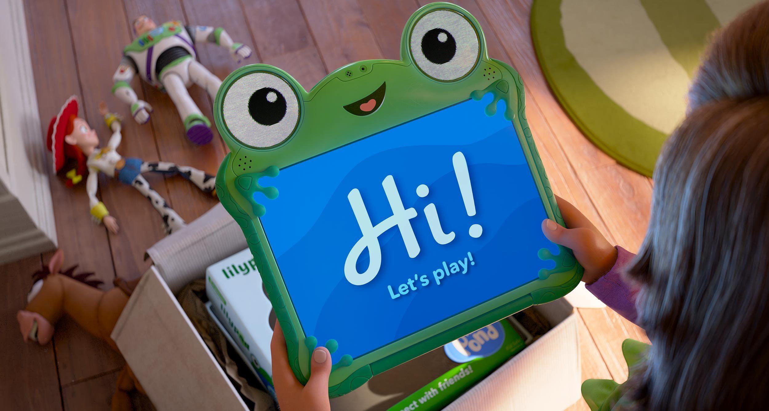 Ni&ntilde;a sostiene una tablet verde con forma de rana que muestra &ldquo;Hi! Let&rsquo;s play!&rdquo; ("&iexcl;Hola! &iexcl;Juguemos!") mientras juguetes est&aacute;n esparcidos en el suelo.