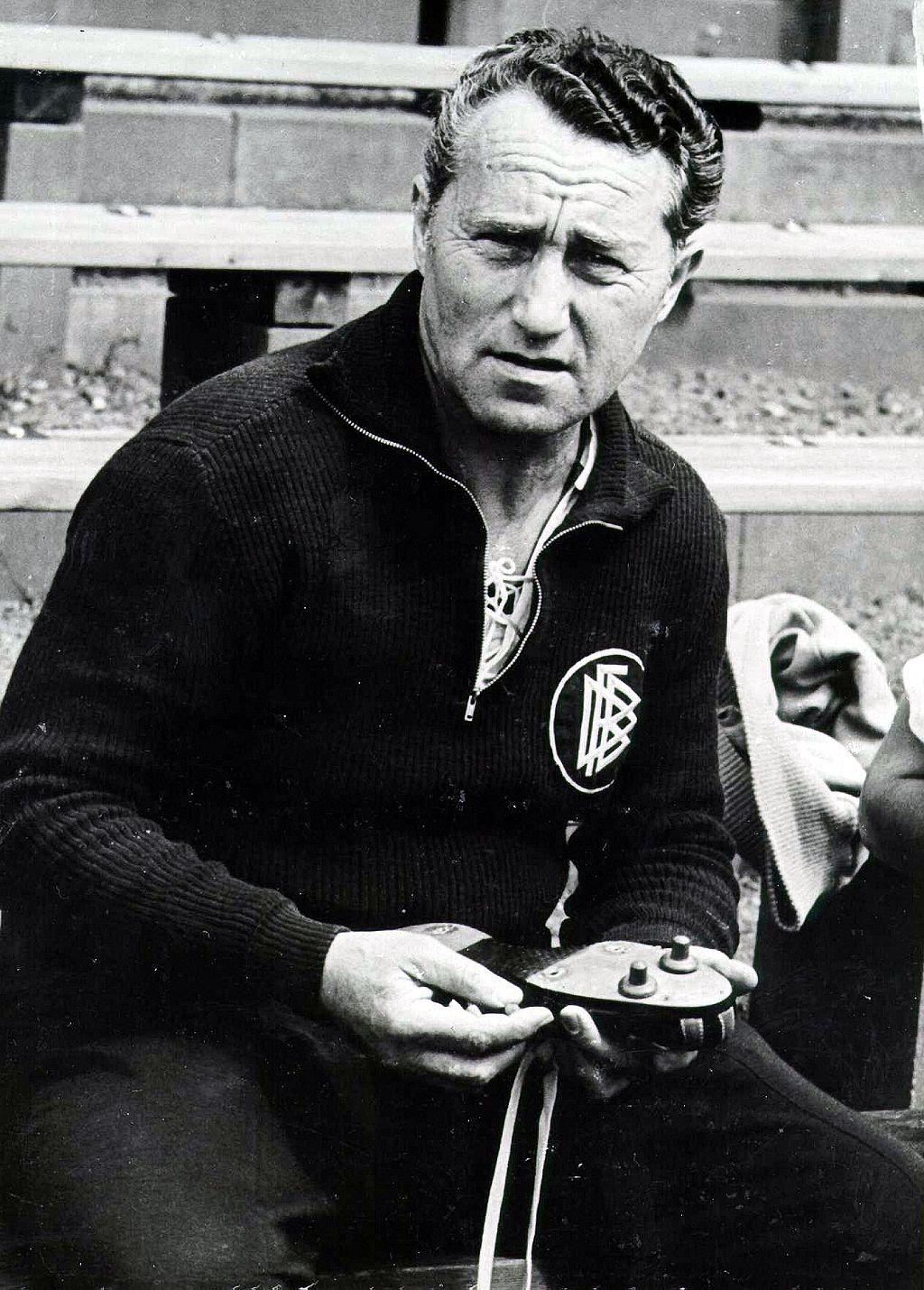 Fotograf&iacute;a en blanco y negro de Adolf Dassler, fundador de la empresa alemana de ropa deportiva Adidas, con ropa oscura y la insignia de la Federaci&oacute;n Alemana de F&uacute;tbol (DFB) a un lado en el pecho, sosteniendo zapatillas de f&uacute;tbol con dos tacos y huecos para fijar m&aacute;s. 
