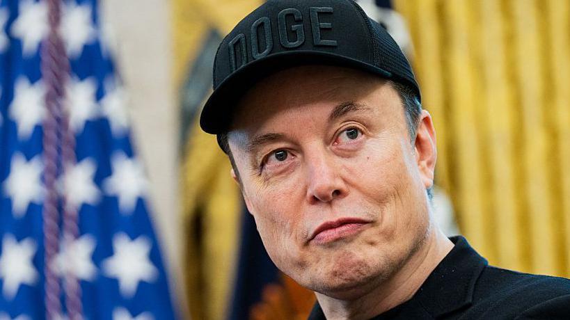 Primer plano de Musk con gorra y la bandera estadounidense de fondo. 