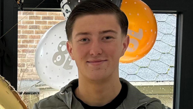 Un joven de 18 a&ntilde;os de pie frente a unos globos naranjas, blancos y negros junto a una ventana. Tiene el pelo corto y oscuro, est&aacute; sonriendo y lleva una chaqueta azul y una camiseta negra debajo.