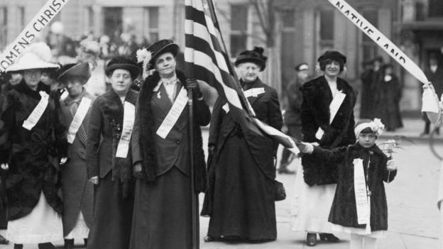 Una foto en blanco y negro de 1909 en el que se ve a mujeres marchando en favor de la prohibici&oacute;n del alcohol en EE.UU. Una en la mitad lleva una bandera estadounidense, la cual es desplegada por una ni&ntilde;a- 