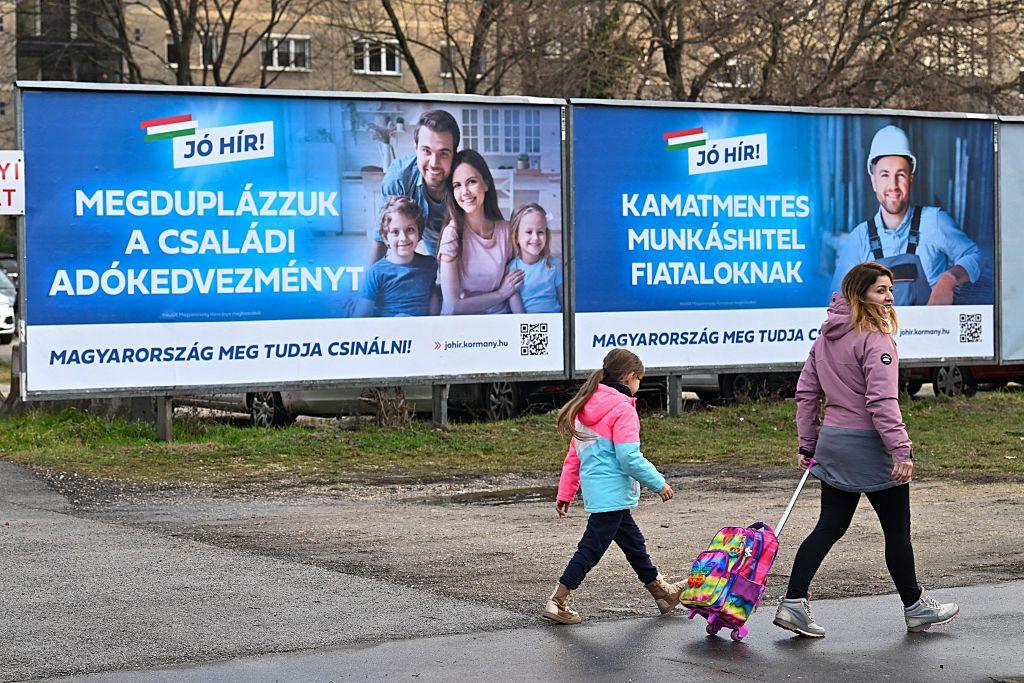 Una madre y su hija caminan por la acera, frente a dos carteles publicitarios. El primero muestra a una familia de cuatro personas con el lema "Duplicamos las ayudas fiscales para familias". El segundo muestra a un trabajador y el lema "Pr&eacute;stamos sin intereses para j&oacute;venes trabajadores".
