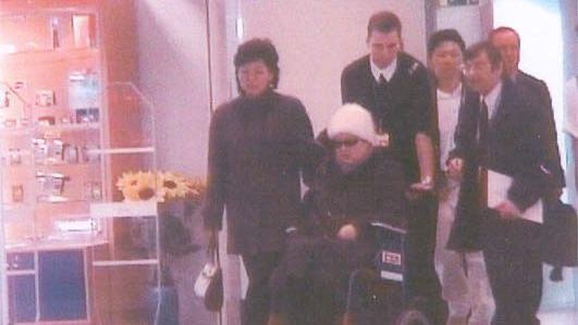 Ko Yong-hui, con gafas de sol y un gorro blanco, est&aacute; sentada en una silla de ruedas que empuja un hombre. Hay varias personas a su alrededor en un hospital de Francia.