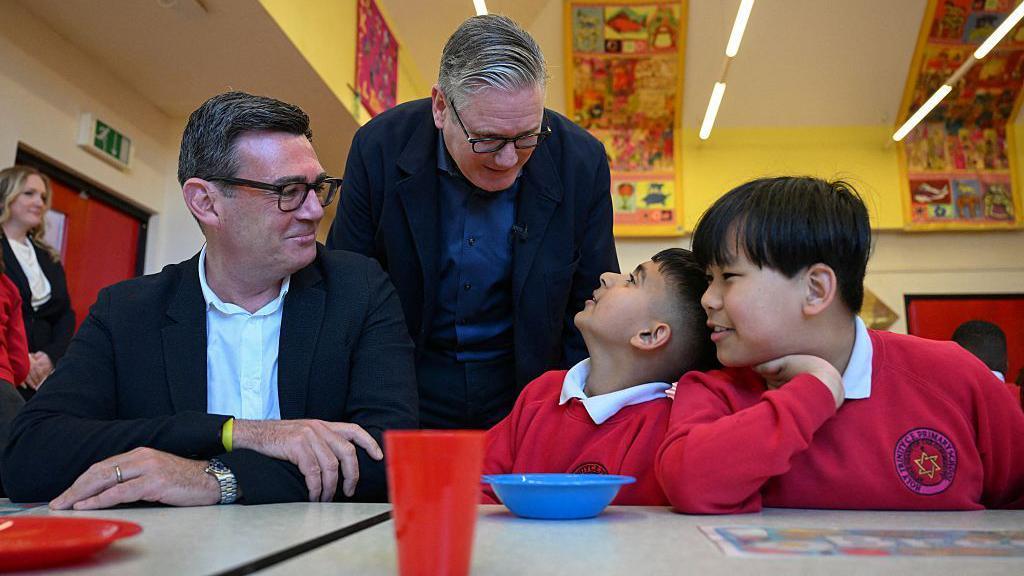 Keir Starmer mira por encima del hombro de Andy Burnham mientras charlan con dos ni&ntilde;os con uniforme escolar durante una visita a un club de desayuno en Gran Manchester, en abril.