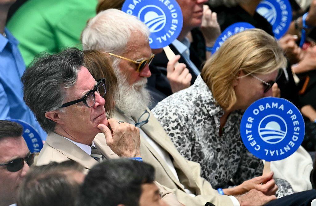 Stephen Colbert y David Letterman siguen el evento rodeados de m&aacute;s personas.  Algunas de ellas portan unos carteles circulares del Obama Presidential Center