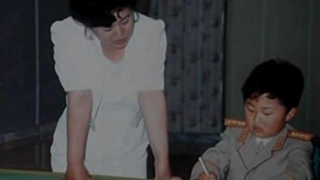 Ko Yong-hui, vestida de blanco, est&aacute; de pie a la izquierda. Un joven Kim Jong-un, vestido con uniforme militar, est&aacute; sentado y escribe con un bol&iacute;grafo. 