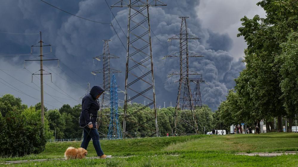 Una mujer pasea su perro por un parque y de fondo se ven las columnas de humo negro de la refiner&iacute;a atacada por Ucrania.