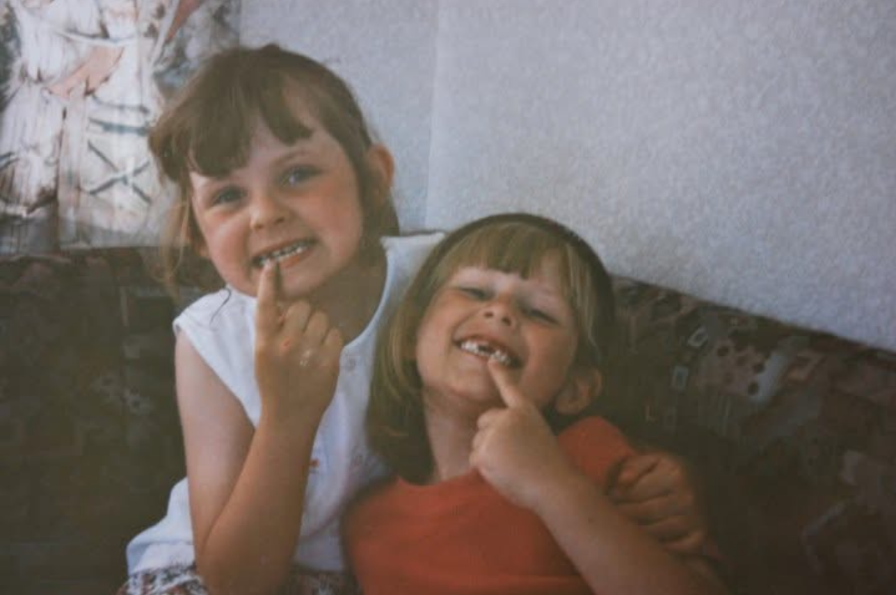 Dos ni&ntilde;as sonriendo a la c&aacute;mara, ambas se&ntilde;alan los huecos dejados por sus dientes faltantes. 