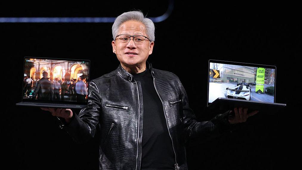 El director ejecutivo de Nvidia, Jensen Huang, sostiene en alto dos computadores port&aacute;tiles sobre un escenario, vistiendo su caracter&iacute;stica chaqueta de cuero negra.