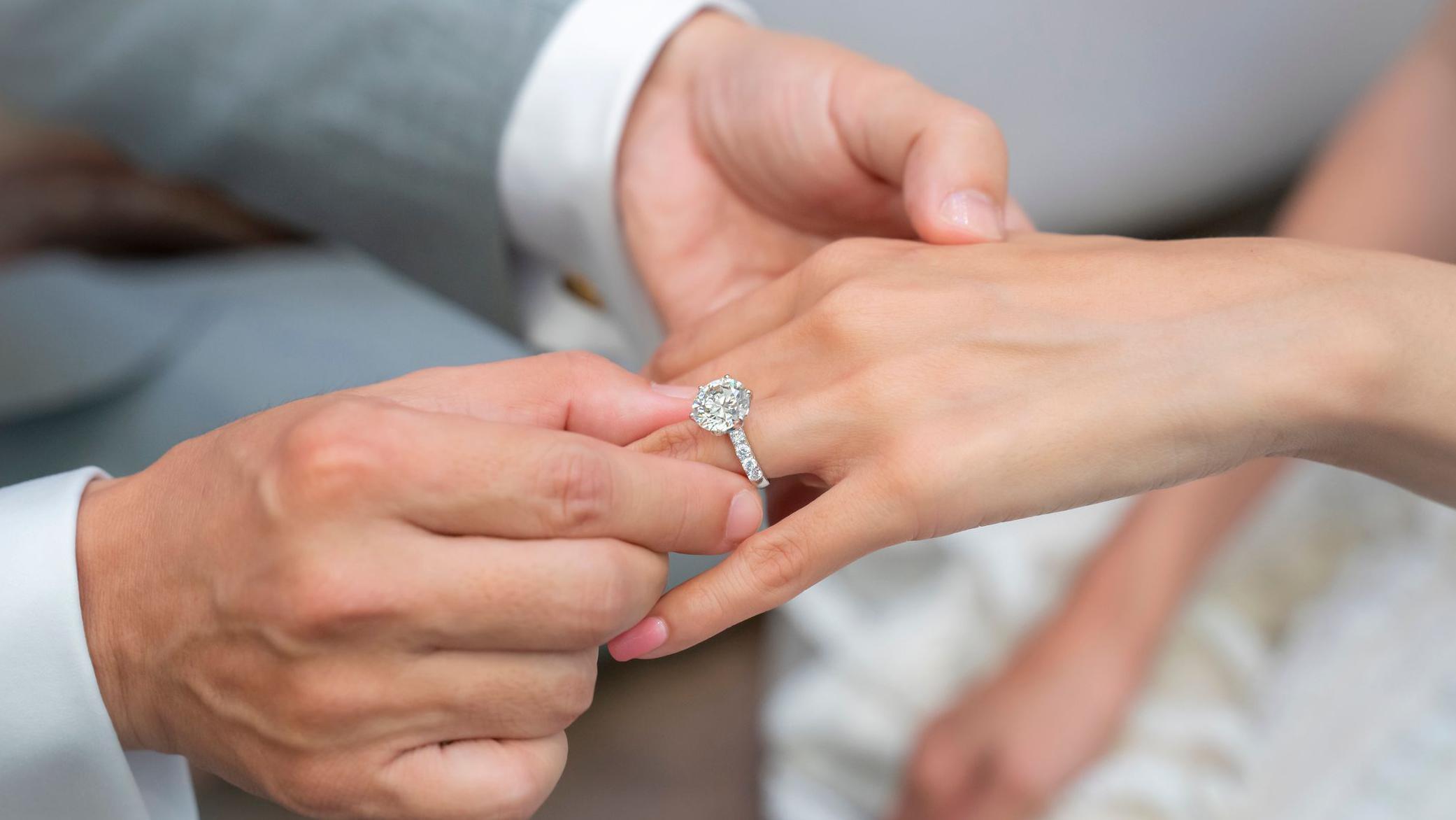 Un hombre coloca un anillo de compromiso con un diamante sint&eacute;tico en la mano de su pareja.