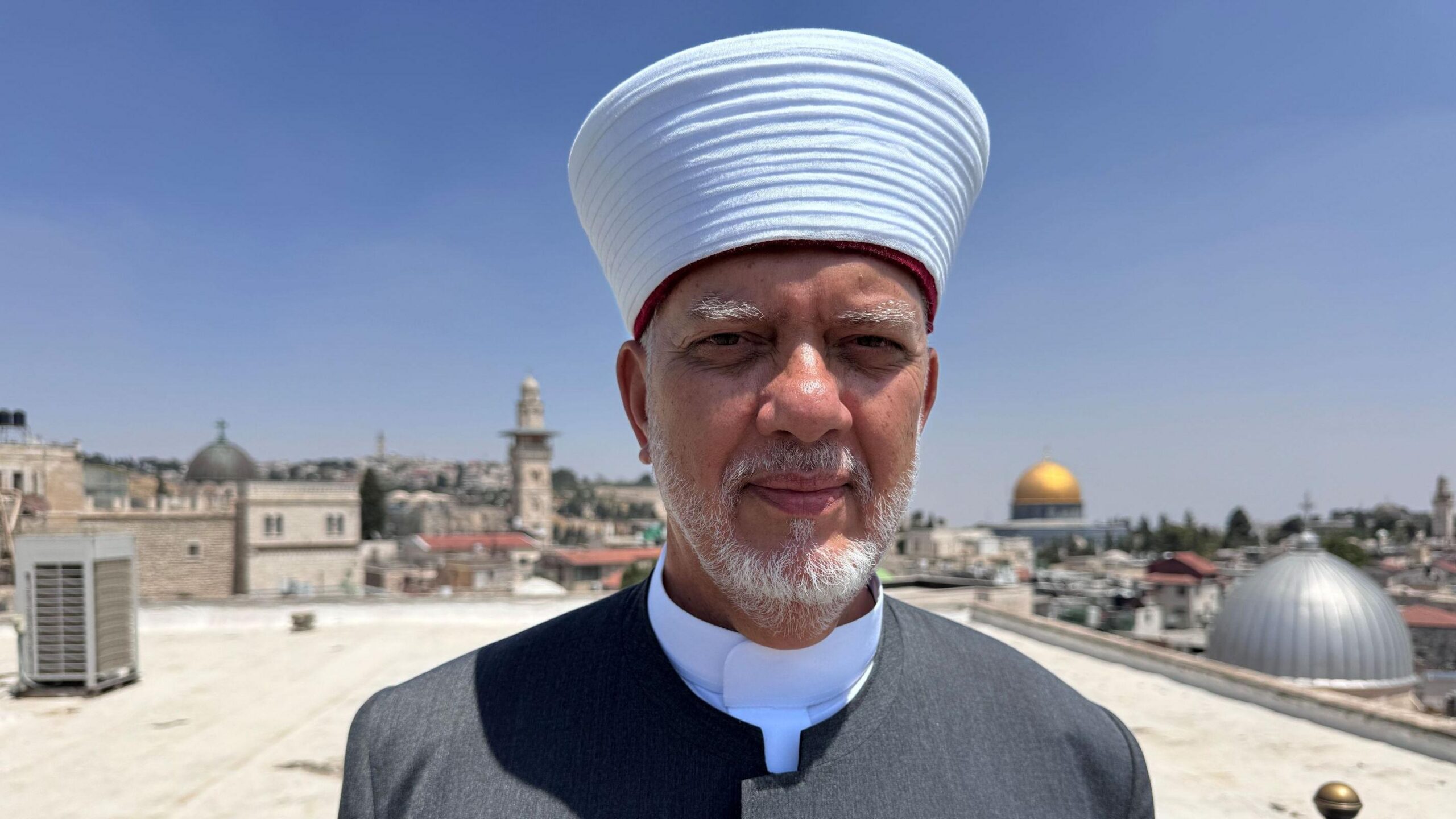 Un hombre con barba y t&uacute;nica gris de pie en una azotea en Jerusal&eacute;n, con la c&uacute;pula dorada de la mezquita de Al Aqsa al fondo.
