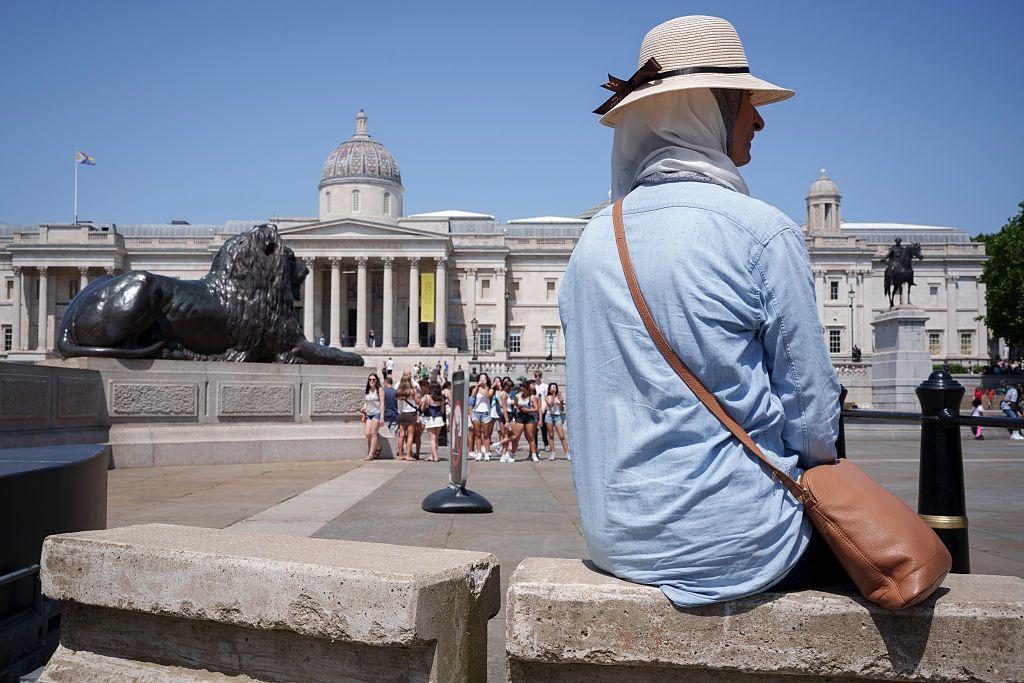 Mujer con camisa delgada y holgada de manga larga, protegida con un pa&ntilde;uelo y un sombrero, est&aacute; sentada frente a la Galer&iacute;a Nacional de Londres. 