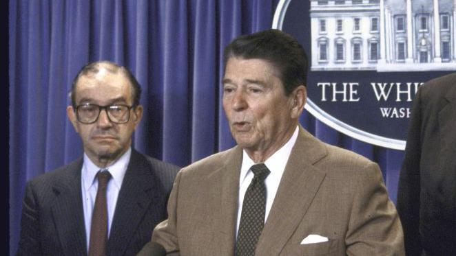 Ronald Reagan y Alan Greenspan en 1987. Est&aacute;n de pie en el podio de la Casa Blanca. El presidente Reagan est&aacute; hablando y Alan Greenspan espera a ser presentado.