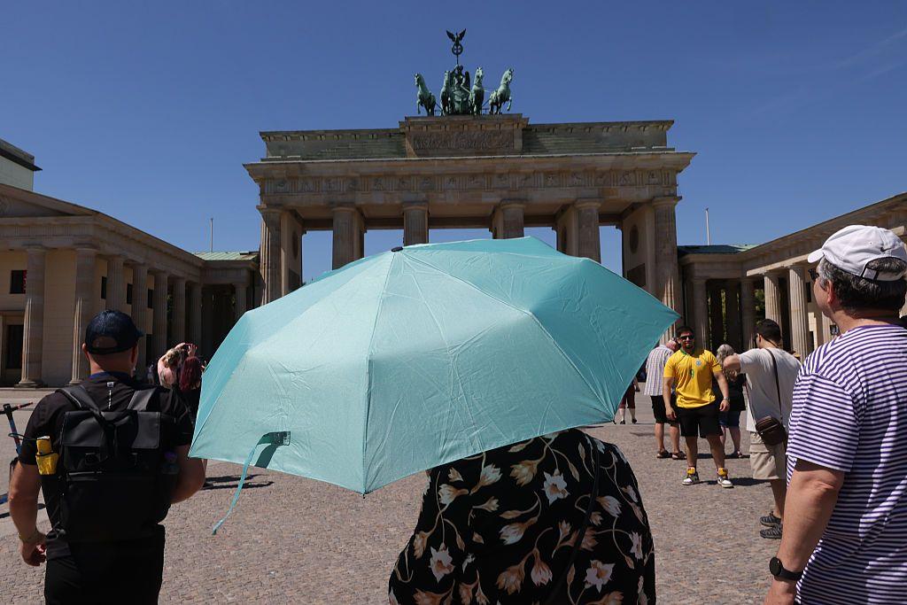 Turistas, uno de ellos bajo una sombrilla, frente a la Puerta de Brandeburgo, en Berl&iacute;n, durante un d&iacute;a soleado.
