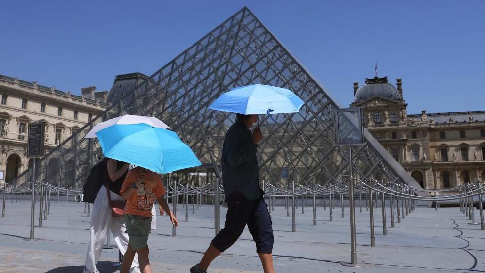 Tres turistas se protegen del sol con sombrillas mientras caminan frente a la Pir&aacute;mide del Museo del Louvre.