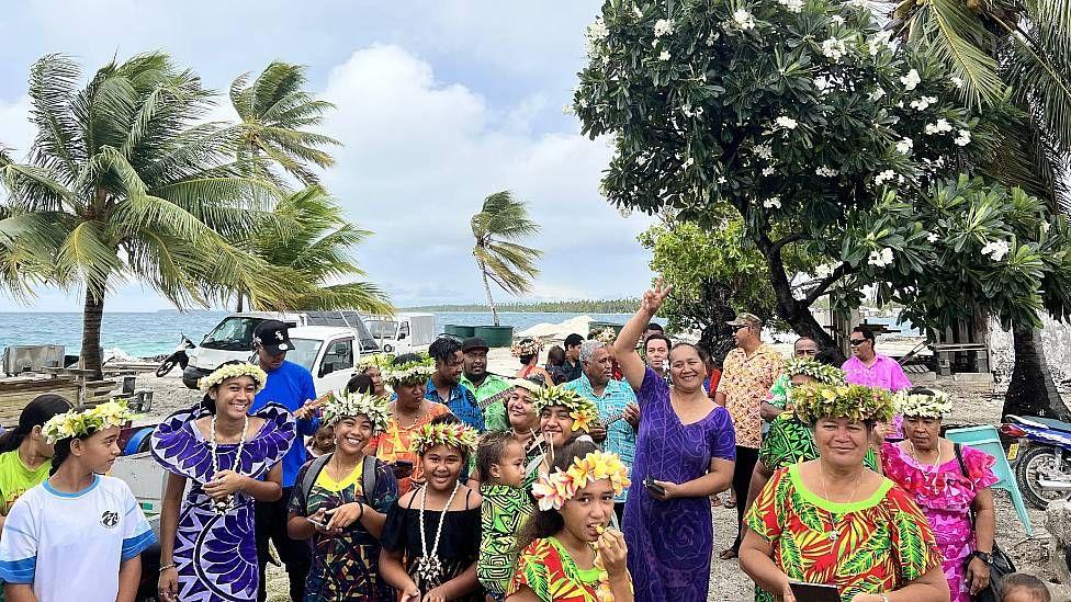 Pobladores de Penrhyn junto al mar sonriendo a la c&aacute;mara. Las mujeres llevan vestidos coloridos y coronas de flores. 