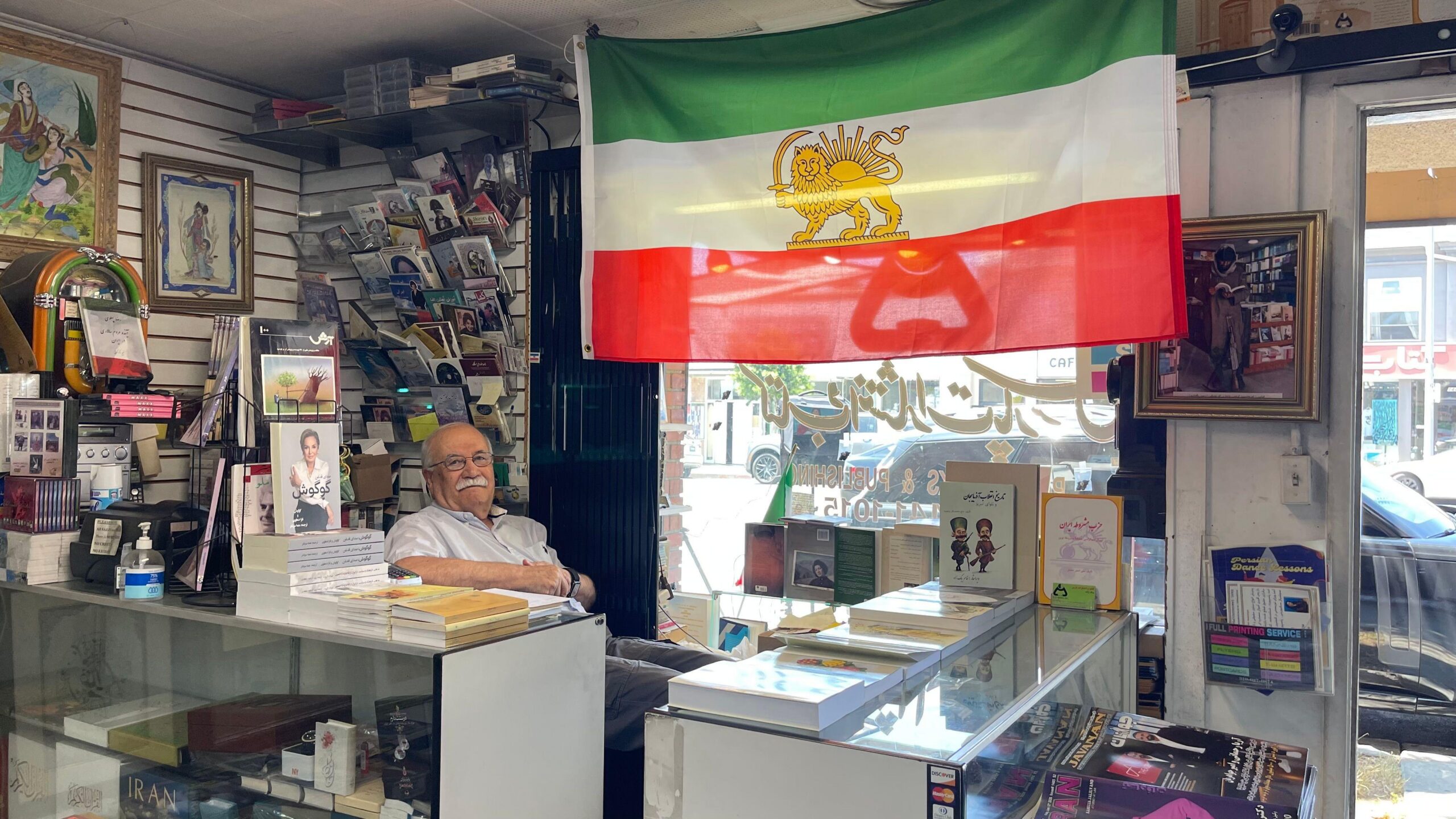 Un miembro de la di&aacute;spora iran&iacute; en Los &Aacute;ngeles mira a c&aacute;mara sentando detr&aacute;s del mostrador de su librer&iacute;a, en la que tiene colgada la bandera previa a la revoluci&oacute;n isl&aacute;mica con el le&oacute;n y el sol, en Teher&aacute;ngeles, Los &Aacute;ngeles, Estados Unidos, el 15 de junio de 2026. (Foto: Leire Ventas)