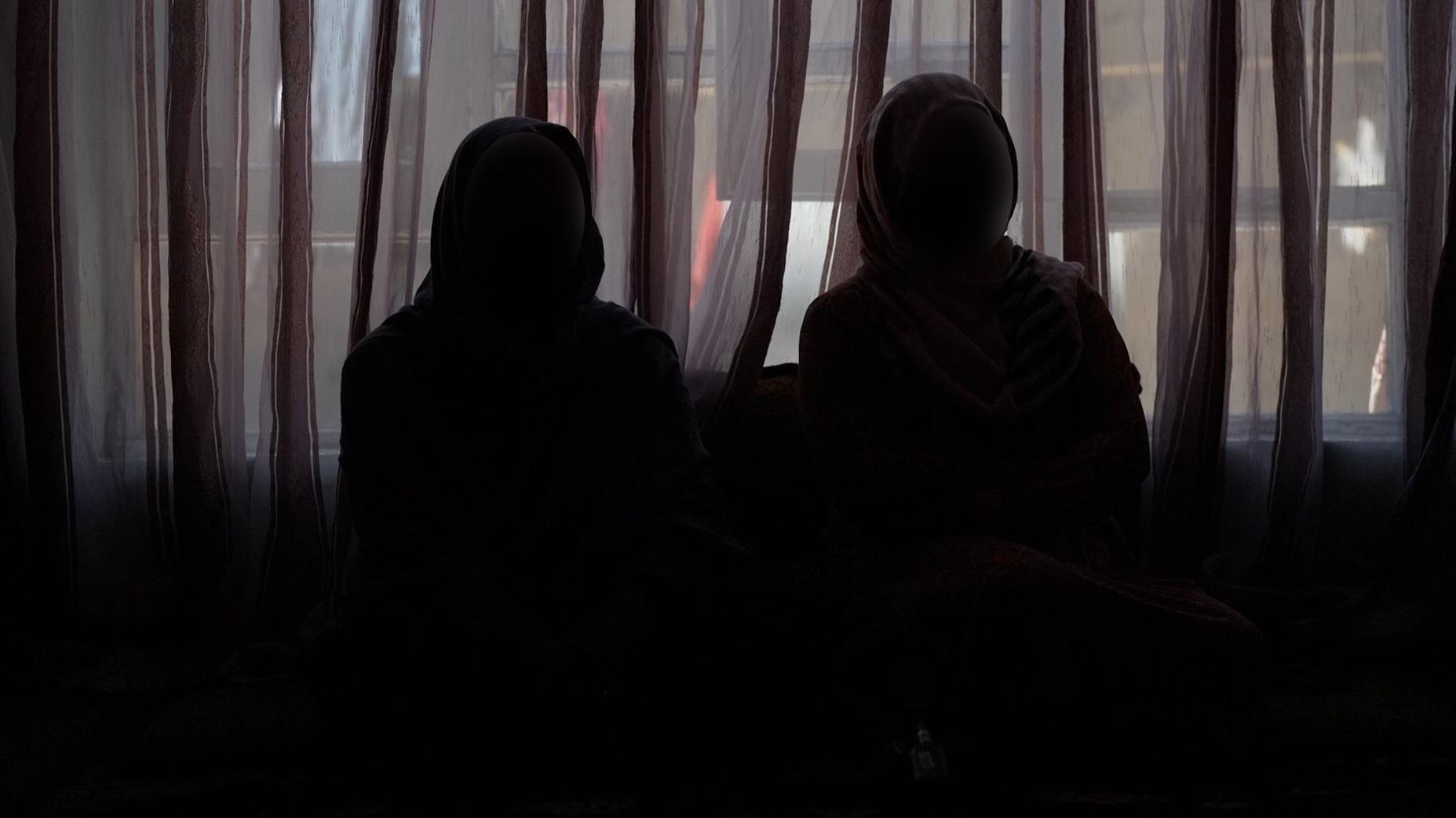 Dos mujeres se sientan en la oscuridad con hiyab y mascarillas para proteger su identidad.
