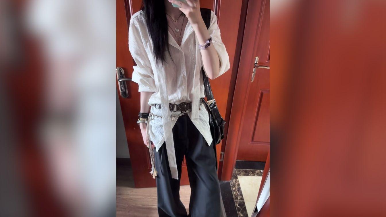 Mujer de pie haci&eacute;ndose un selfie frente a un espejo con una pared roja en el fondo, vestida con pantalones negros y camisa blanca con flecos largos