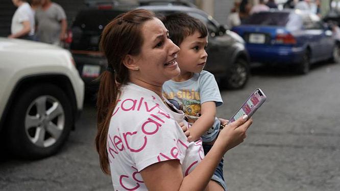 Una mujer con cara de angustia carga a un ni&ntilde;o peque&ntilde;o con una mano mientras sostiene su celular con la otra mano en una calle de Caracas, luego del doble terremoto del 24 de junio.