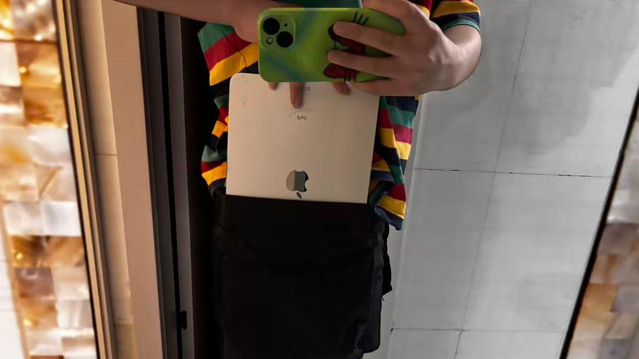 Una mujer vestida con ropa masculina de talla grande sostiene un tel&eacute;fono m&oacute;vil en la mano izquierda, toma una foto mientras guarda una tableta port&aacute;til en el bolsillo con la otra mano. Su camiseta es multicolor y lleva pantalones negros