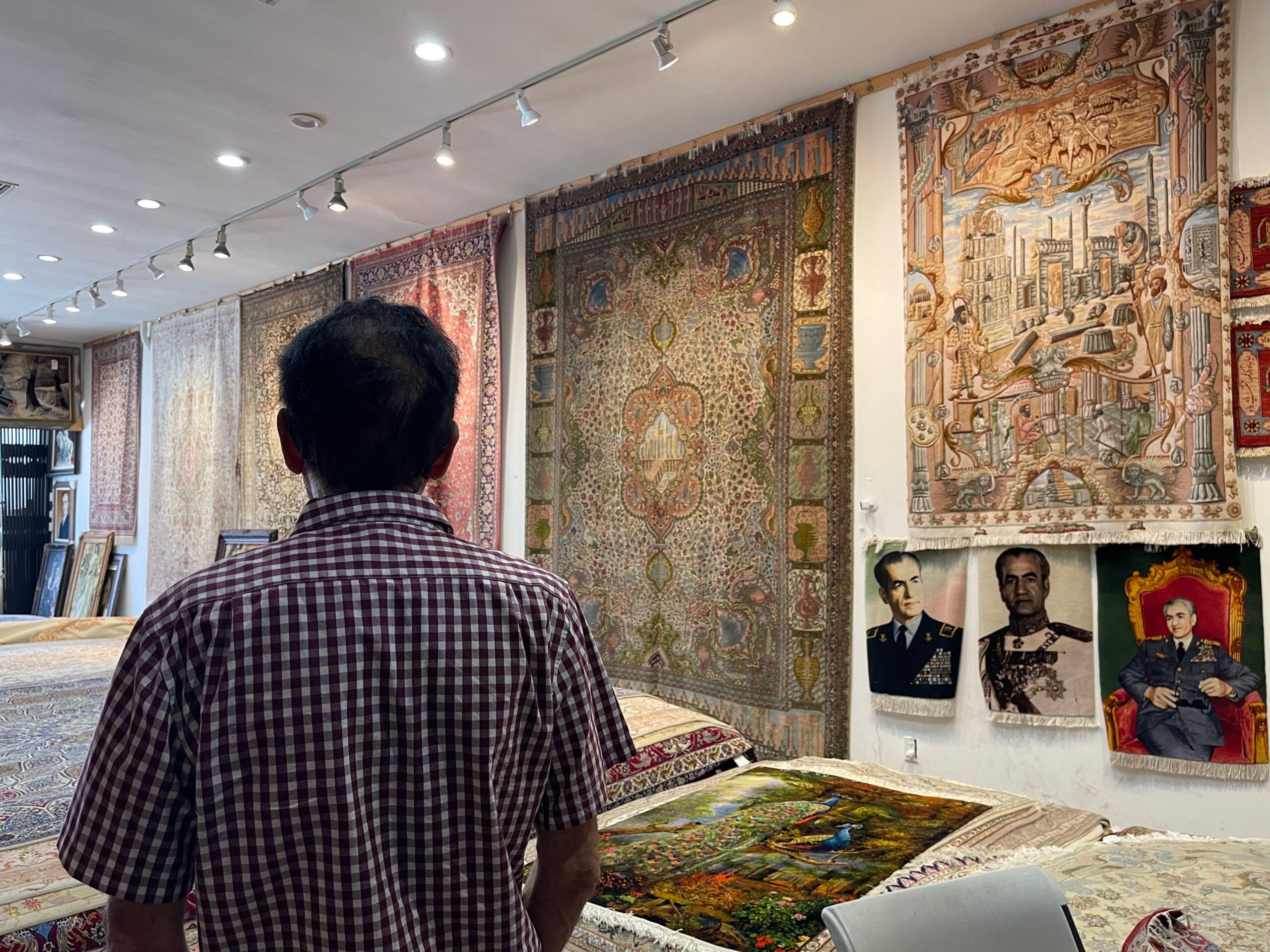 Eric Saddith de espaldas en su tienda alfombras de Teher&aacute;ngeles, con alfombras colgadas de las paredes y tapices con el retrato del &uacute;ltimo sha de Ir&aacute;n, Mohamed Reza Pahlavi, el 15 de junio de 2026. (Foto: Leire Ventas)
