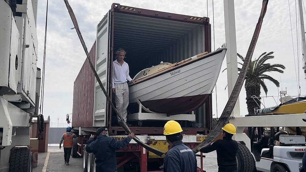 Robinson junto a su bote en un contenedor cargado en la parte trasera de un cami&oacute;n. En el suelo hay tres operarios que tendr&aacute;n a su cargo descargar el bote.