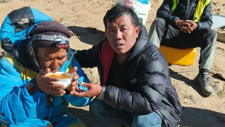 Un hombre con una chaqueta de escalada azul bebiendo una sopa naranja de un cuenco, siendo asistido por un hombre con una chaqueta acolchada negra.