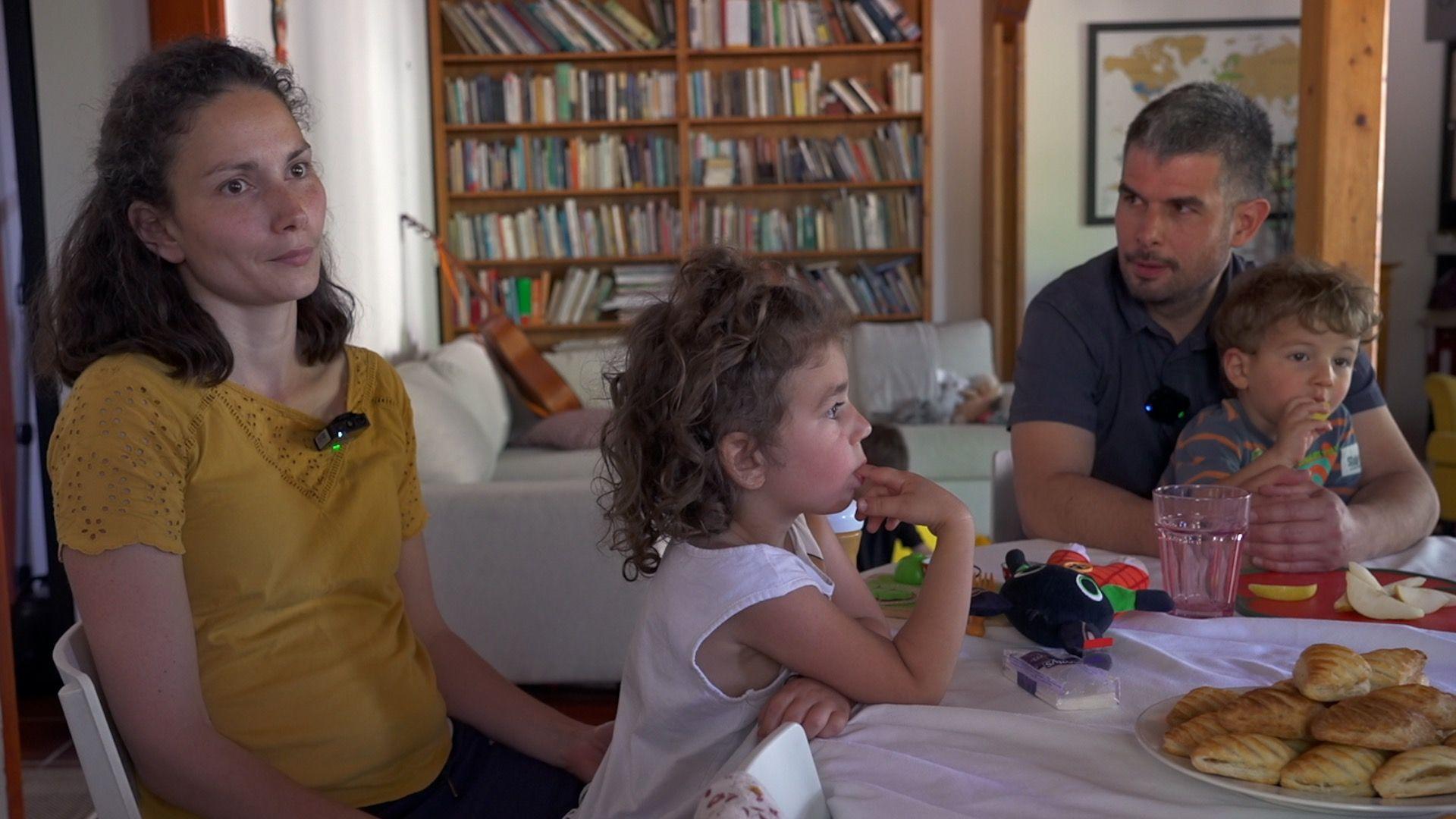 Una mujer y un hombre est&aacute;n sentados alrededor de una mesa, cada uno con dos ni&ntilde;os peque&ntilde;os en su regazo. Sobre la mesa hay pasteles y juguetes. Al fondo se ve una estanter&iacute;a, un sof&aacute; y, encima de este, una guitarra.