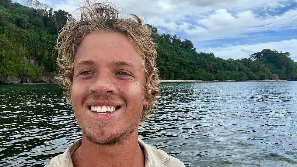 Tom Robinson sonriendo a la c&aacute;mara. Detr&aacute;s se ve el mar y la vegetaci&oacute;n en la cosa de una isla del Pac&iacute;fic.